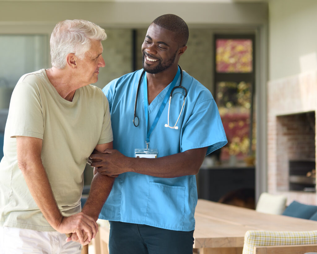 Male Nurse Visiting Senior Man Using Walking Stick With Mobility Issues At Home